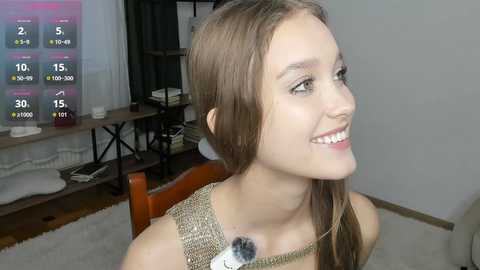 Video of a smiling young woman with fair skin and straight brown hair, wearing a beige sweater, sitting on a wooden chair in a modern, cluttered living room.