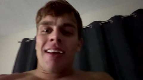 A video of a shirtless, light-skinned teenage boy with light brown hair, wearing a playful expression, leaning against a dark shower curtain in a dimly lit bathroom.