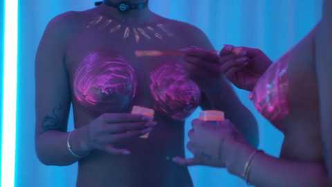 Video of a topless, muscular Black woman with painted pink nipples and tribal markings on her chest, holding lit candles, against a blue-lit background.