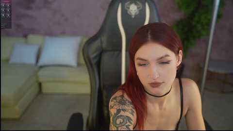Media: Video of a pale-skinned, red-haired woman with tattoos on her left arm, wearing a black choker, sitting in a gaming chair in a modern living room with a beige sofa and a green plant.