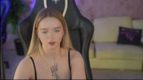 Video of a young, fair-skinned woman with long blonde hair, wearing a black tank top, seated in a black gaming chair. Background features a beige couch and a pink wall.
