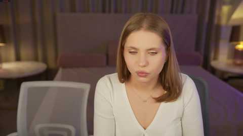 Video of a Caucasian woman with straight brown hair, wearing a white blouse, sitting at a modern, transparent desk in a dimly-lit bedroom with beige walls and two bedside lamps.