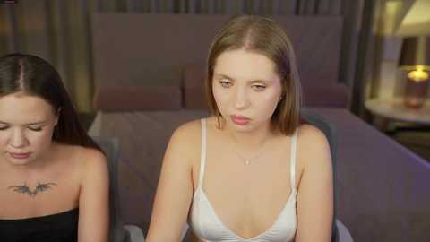 Media: Video of two young women, one with short brown hair and a white spaghetti strap top, and another with long dark hair in a black strapless top, sitting in a dimly lit bedroom.