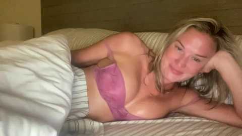 Video of a blonde woman lying on a bed in a light-colored, striped bedspread, wearing a purple lace bralette and gray high-waisted pants. She has fair skin and is relaxed, with a thoughtful expression.