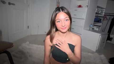 Media: A video of a young, light-skinned woman with long, straight brown hair, wearing a black polka-dot bandeau top, sitting on a white shag rug in a modern, white-walled bedroom.