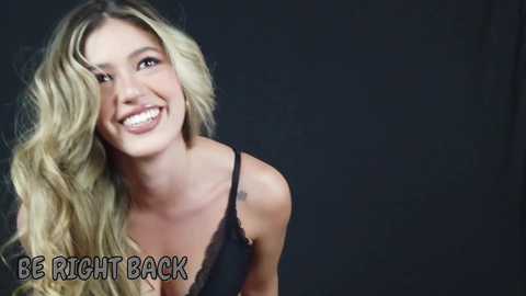 Video of a smiling, fair-skinned blonde woman with wavy hair, wearing a black lace-trimmed bra, against a dark background. Text in the bottom left reads, \"BE RIGHT BACK.\