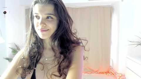 Video of a young woman with long, wavy brown hair and light skin, smiling, wearing a sheer top, in a brightly lit room with a beige curtain and plants in the background.