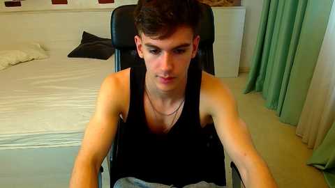 Video of a young, slim, Caucasian man with short dark hair, wearing a black tank top, sitting in a wheelchair in a modern, minimalist bedroom with white walls, beige curtains, and a neatly made bed.