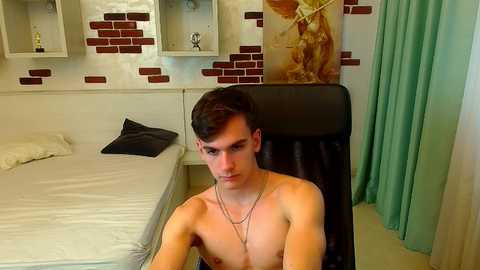 Video of a shirtless, young, fair-skinned man with short dark hair sitting on a black chair in a modern bedroom with white brick wall art, a bed, and teal curtains.