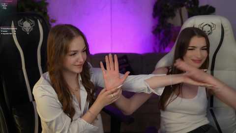 Video of two young women, one with long brown hair, the other with long black hair, interacting playfully with virtual reality controllers in a cozy, dimly lit living room.
