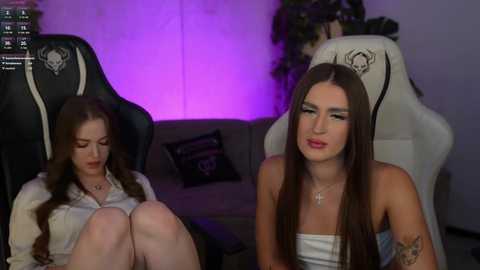Media: Video of two young women in modern gaming chairs, one with long brunette hair, wearing a white top, the other with straight brown hair, wearing a strapless top. Purple lighting and a gray couch in the background.