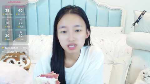 Video of a young Asian woman with long black hair, wearing a white shirt, sitting on a white bed. A digital overlay displays her social media stats and a virtual dog.