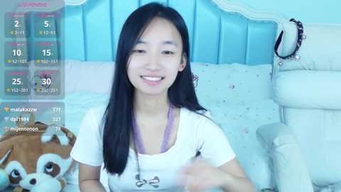 A video of a smiling young Asian woman with long black hair, wearing a white T-shirt, sitting in a light blue room with a plush dog toy, digital stats overlay, and a choker necklace.