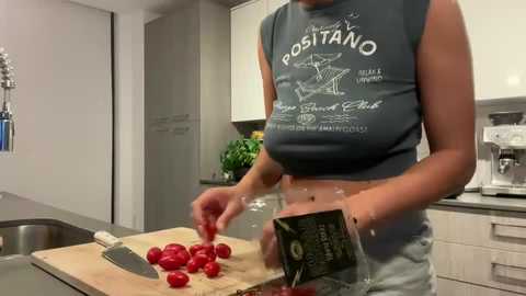 Video of a woman in a modern kitchen, wearing a blue sleeveless crop top with \"Positano\" text, cutting cherry tomatoes on a wooden cutting board.