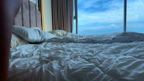 Media: Video of a messy bed with white sheets and pillows, next to a large window showing a blue sky with scattered clouds, creating a serene and relaxed atmosphere.