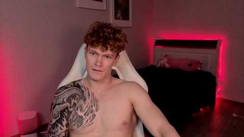 Video of a shirtless, pale-skinned, curly-haired man with a large black and gray tattoo on his left shoulder, sitting in a black chair under red LED lighting.