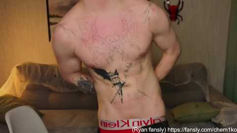 Video of a shirtless, muscular, light-skinned man with tattoos on his chest and abdomen, wearing Calvin Klein underwear, standing in a dimly lit living room with a grey couch and a red wall.