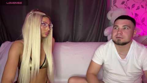 A video of a blonde woman in a gold bikini and a bearded man in a white T-shirt, sitting on a couch against a dark curtain backdrop, illuminated by pink light.