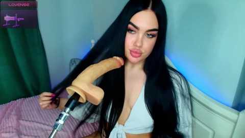 Video of a young woman with long black hair, fair skin, and dark makeup, wearing a white bra, holding a large, lifelike dildo. Bedroom background with blue lights, bed, and green curtains.