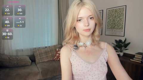 Media: Video of a young woman with fair skin, long blonde hair, wearing a light pink dress with floral patterns, sitting in a cozy living room with beige furniture, framed artwork, and a digital camera overlay.