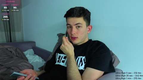 Video of a young man with short dark hair, wearing a black shirt with white text, holding a phone, lying on a bed with gray sheets, against a light blue wall.
