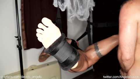 A video shows a muscular, shirtless man's tattooed arm with a black strap tied around his wrist, suspended mid-air. Background includes white walls, a black chair, and a hanging white garment. URL watermarks are visible.