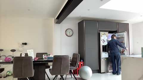 A modern, minimalist kitchen with a white wall, a large black fridge, and a man in a blue shirt opening it. The room features a brown dining table, chairs, and a round table with a lamp.