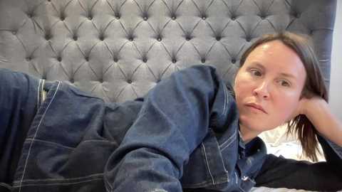 Media: Video of a Caucasian woman with light skin, brown hair, and blue eyes, lying on her side on a bed with a gray tufted headboard. She wears a dark denim jacket and black shirt, looking relaxed and contemplative.