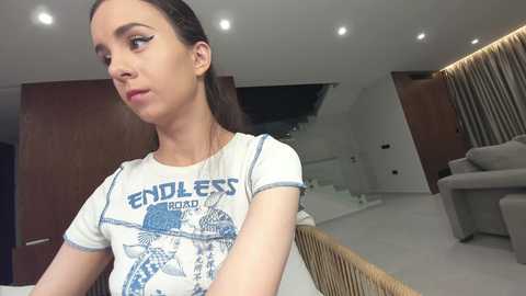 Video of a slender Caucasian woman with dark hair, wearing a white \"Endless\" t-shirt, standing in a modern, minimalist living room with white walls and wooden accents.