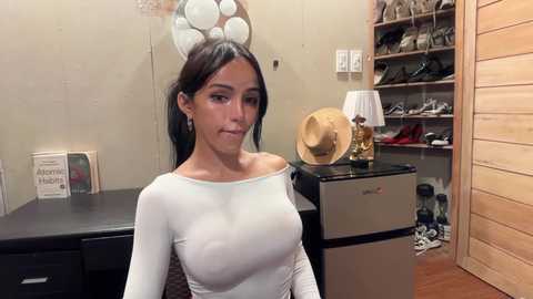 Video of a young woman with long dark hair, wearing a tight, off-the-shoulder white top, standing in a cluttered room with beige walls, a small refrigerator, and a bookshelf.