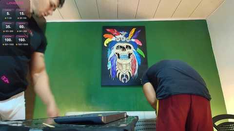 Video of two men in a studio, one with a black shirt, the other with red shorts, interacting with a green wall featuring a vibrant, colorful feathered headdress painting.