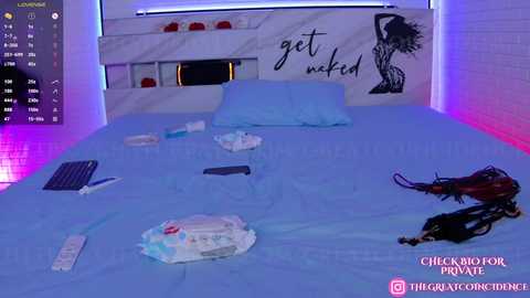 Video of a modern bedroom with a blue bed, white headboard, and a \"get naked\" mural. On the bed, there are a diaper, phone, and a pair of black lingerie.
