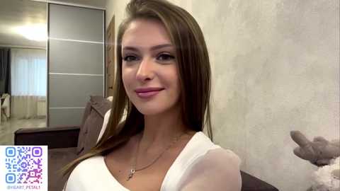 A video of a smiling young woman with straight brown hair, wearing a white top, standing in a modern, beige-tiled room with a plush grey armchair and a closet in the background.