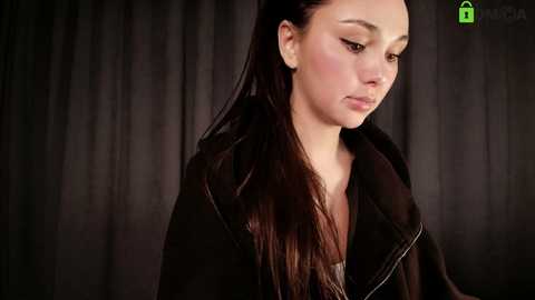 Video of a young Asian woman with long, straight black hair, wearing a black leather jacket, standing against dark, textured curtain backdrop. She appears introspective and serious.