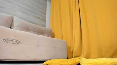 Media: Video of a modern bedroom featuring a beige upholstered bed with silver handles, paired with vibrant mustard-yellow curtains. The background has light wooden paneling, and the floor is covered with dark wood. The scene exudes a cozy, contemporary vibe.