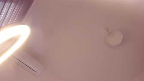 A close-up video of a nude, pale-skinned woman with a small, round nipple and a faint areola. The background includes a blurred, warm-toned light source, likely a lamp, casting a soft glow on the skin.