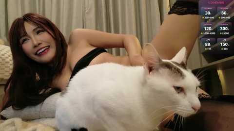 Video of an Asian woman with long brown hair, smiling, lying on a bed, wearing black lingerie, with a white cat on her stomach.