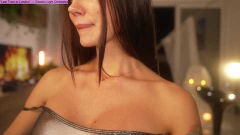 Media: A close-up video of a smiling woman with straight, shoulder-length brown hair, wearing a strapless silver sequin dress. The background features a softly lit room with blurred furniture and warm lighting.