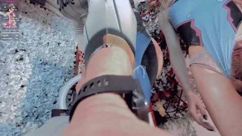 Video of two women in a playful, slightly chaotic setting. One woman, in a blue and black athletic top, is restrained by a black harness. The other, in a light blue top, holds her hands up. Background is messy with scattered objects.
