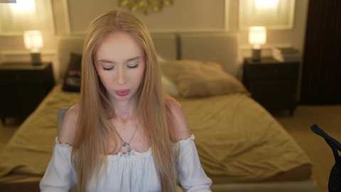 Video of a blonde woman with long hair, wearing a white off-shoulder top, looking down in a dimly-lit bedroom with beige bedding, two bedside lamps, and a dark wardrobe.