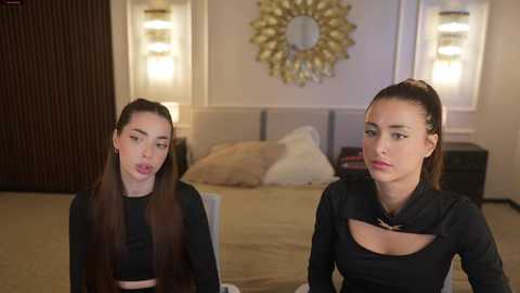 Video of two women in a modern bedroom; one with long hair, the other with a high ponytail, both wearing black tops, looking bored.