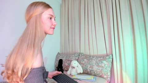 A video of a young woman with long, straight blonde hair in a headband, wearing a gray off-the-shoulder top, typing on a laptop in a cozy, pastel-colored room with floral-patterned cushions and a stuffed animal.
