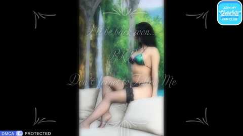 Video of a woman with long black hair in a green bikini, sitting on a cream sofa, with a lush forest backdrop.