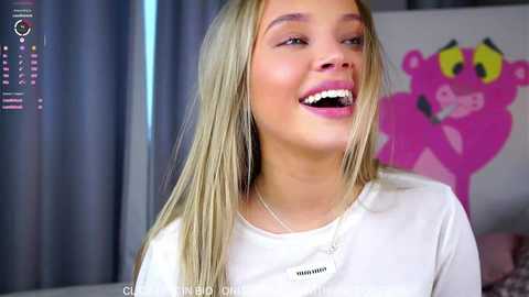 Video of a young, smiling Caucasian woman with long blonde hair, wearing a white shirt, in a colorful room with a pink bear poster.