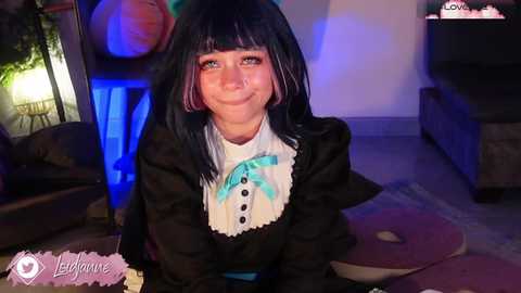 Video of a young woman with long black hair and blue streaks, dressed in a black maid outfit with a white apron and bow, smiling in a dimly lit living room.