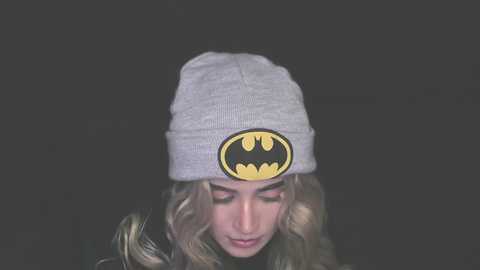 Video of a young woman with long, wavy blonde hair, wearing a gray beanie with a Batman logo, set against a dark, solid background. She has a contemplative expression.