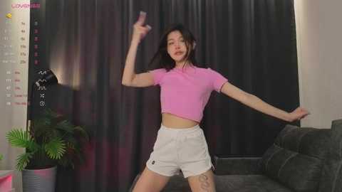Video of a slender Asian woman with medium-length black hair, wearing a pink crop top and white shorts, dancing energetically in a room with dark curtains, green potted plant, and a dark couch.