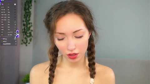 Video of a young woman with fair skin and brown hair in twin braids, wearing a beige top, looking down in a modern, minimally decorated room with a green plant and a computer screen displaying a virtual reality game.
