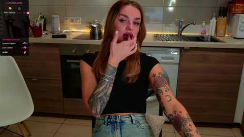 Media: Video of a tattooed woman with long, auburn hair, wearing a black crop top and jeans, eating a burger in a modern kitchen with wooden cabinets and stainless steel appliances.