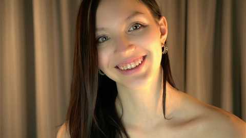 Video of a smiling, fair-skinned woman with long, dark hair, wearing a strapless top, set against beige curtains.